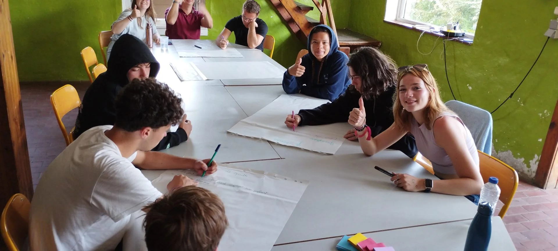 bootcamp_2023_smile_school Plusieurs élèves sont réunis autour d'une table dans une salle aux murs verts. Ils travaillent ensemble en écrivant sur des grandes feuilles blanches. Plusieurs d'entre eux regardent l'appareil photo et lèvent leur pouce.