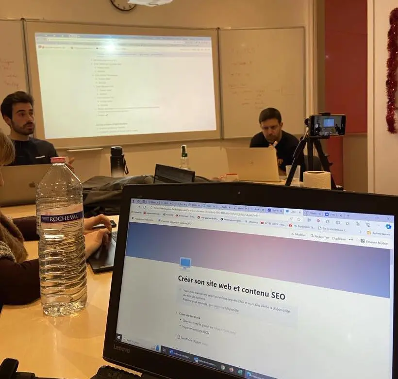 Salle de classe avec le rétroprojecteur allumé. Plusieurs adultes sont réunis autour d'un table et discutent. Au premier plan se trouve un écran d'ordinateur affichant un titre "Créer son site web et contenu SEO".