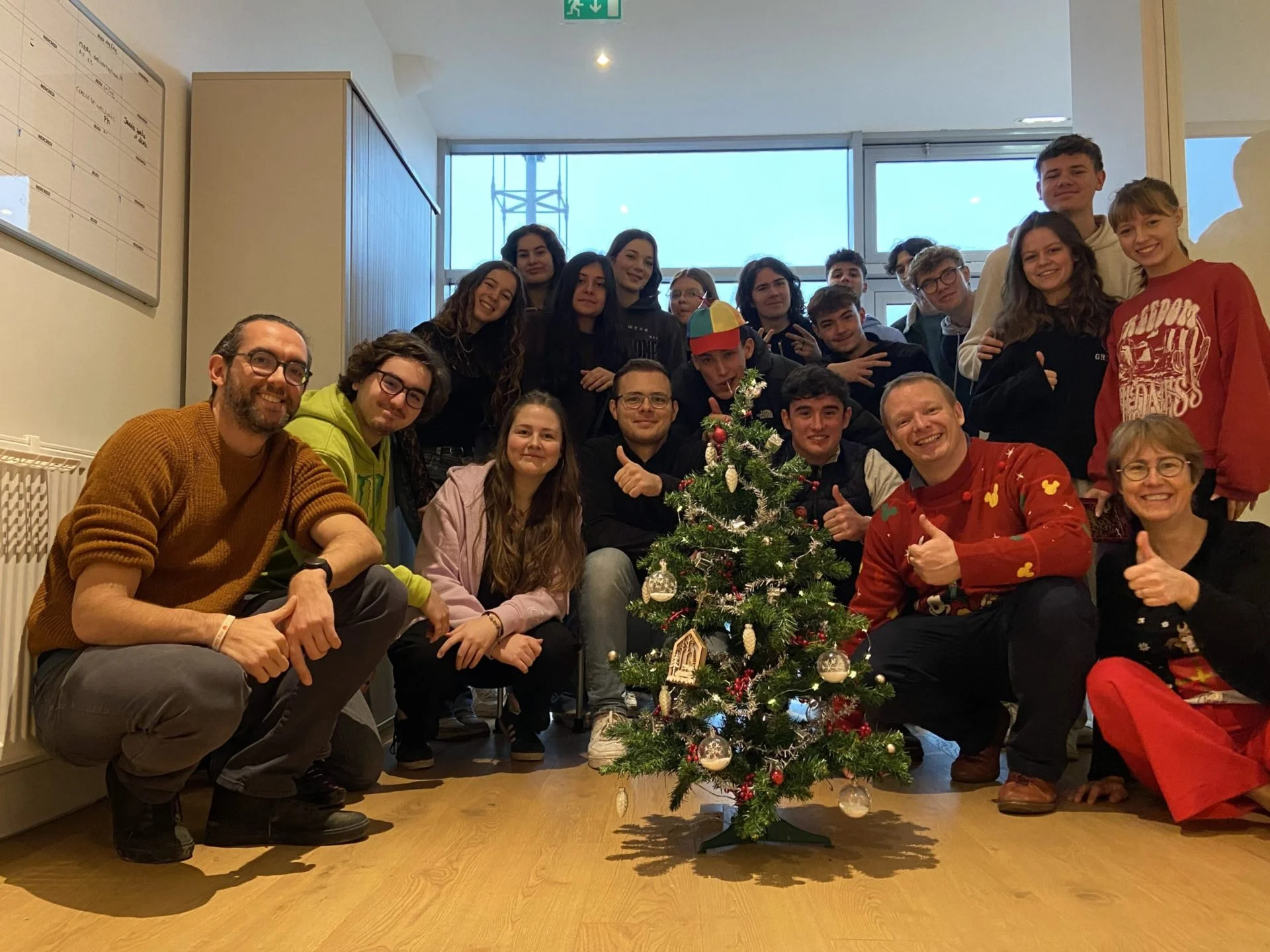 Un groupe d'étudiants et professeurs posent derrière un petit arbre de noël. Ils sourient et certains ont les pouces levés.